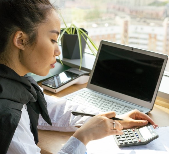 Mulher sentada à frente de um portátil a segurar numa caneta enquanto usa uma calculadora